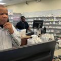 A man answers a phone at a pharmacy counter while another man works on orders at a counter behind him.