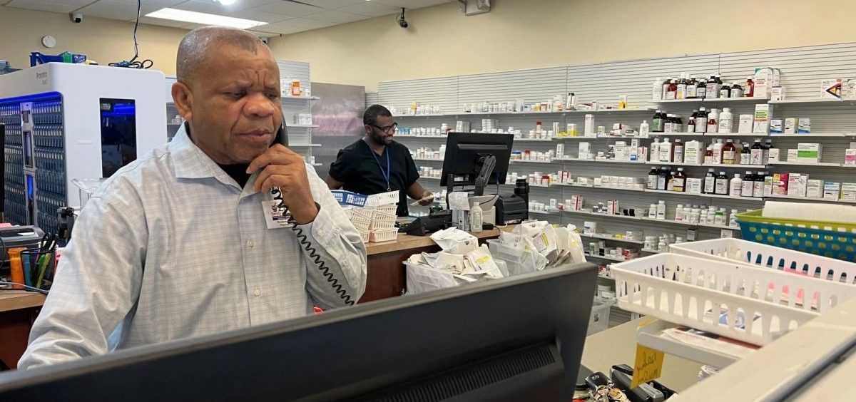 A man answers a phone at a pharmacy counter while another man works on orders at a counter behind him.