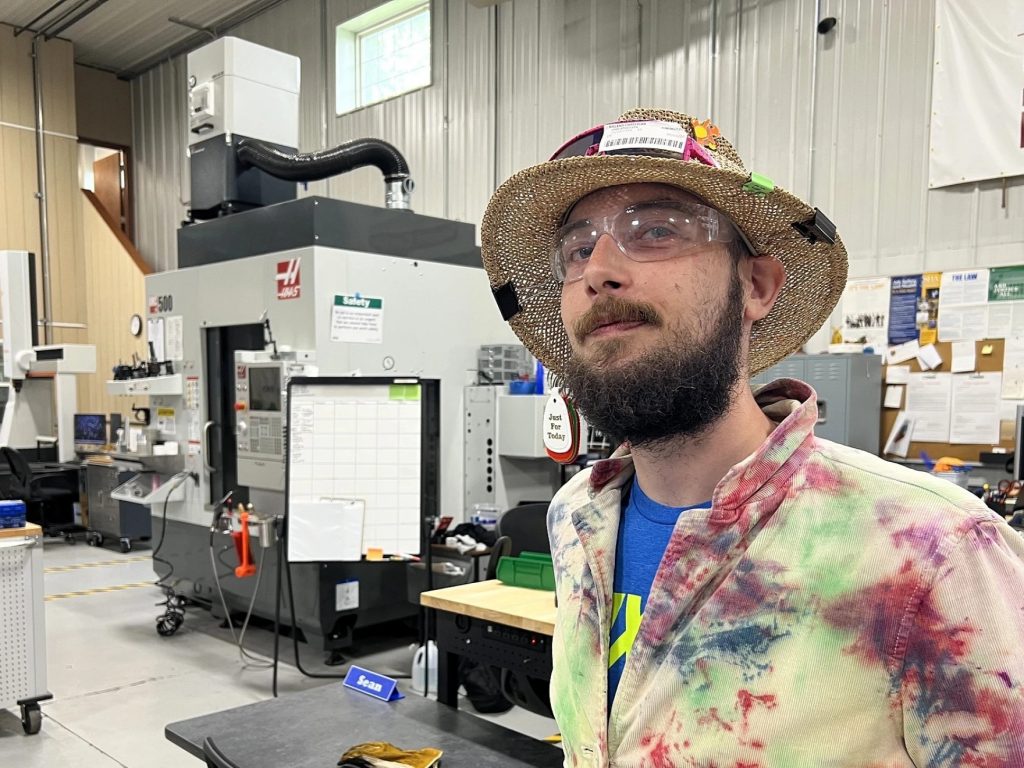 A man in a hat stands in a workshop with large equipment used in manufacturing.