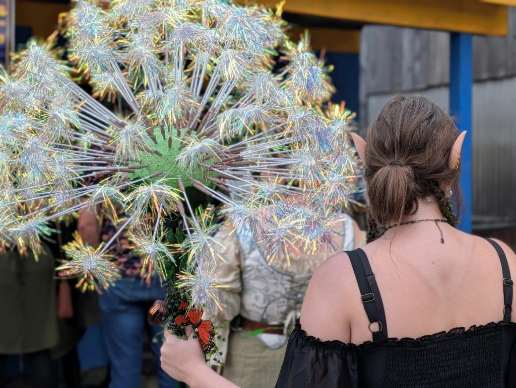 A person with elf ears holds a giant artificial flower arrangement with the stems covered in butterflies.