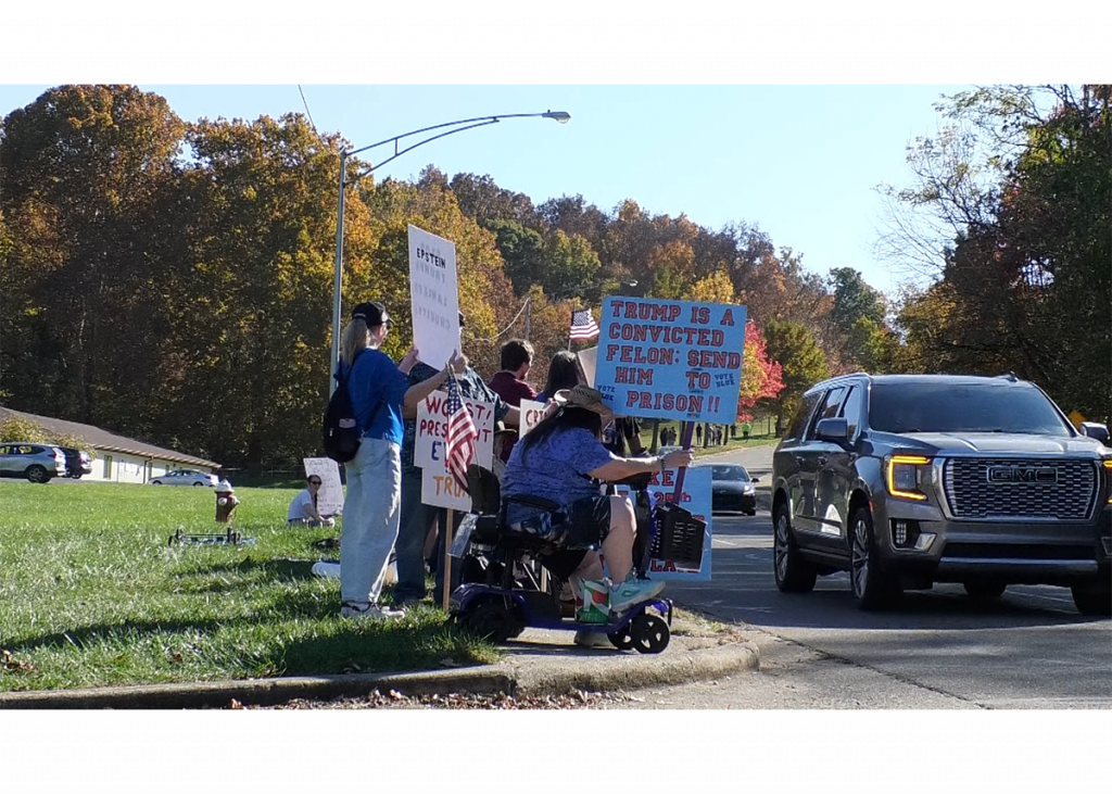 Several protestors hold up signs along the sidewalk of University Drive in Chillicothe, Ohio. Two cars are passing them by.